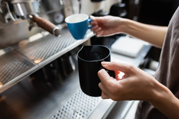Vista ritagliata del barista che tiene la brocca di latte e la tazza offuscata vicino alla macchina del caffè nel caffè — Foto stock