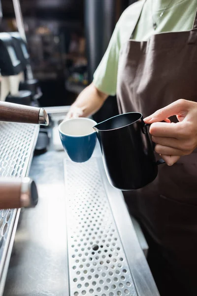 Vista recortada de barista sosteniendo jarra y taza cerca de la máquina de café en la cafetería - foto de stock