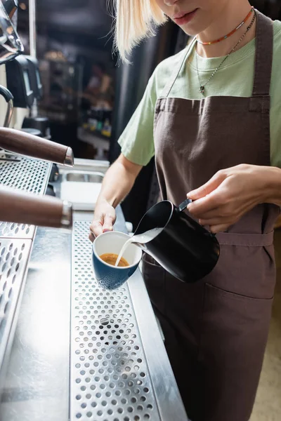 Ausgeschnittene Ansicht von Barista im Schürze gießt Milch in Tasse Kaffee in der Nähe Kaffeemaschine im Café — Stockfoto