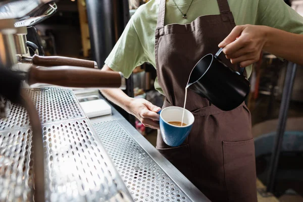 Vista cortada de barista em avental derramando leite de jarro em xícara no café — Fotografia de Stock