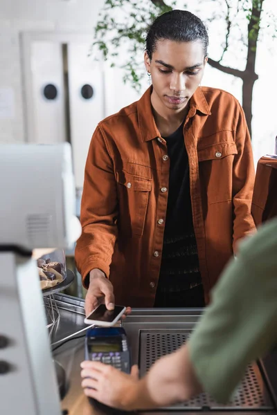 Cliente afroamericano pagando con smartphone cerca de barista borrosa en cafetería - foto de stock