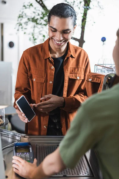 Cliente afroamericano sorridente che punta con smartphone barista quasi sfocato nel caffè — Foto stock