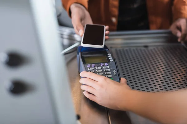 Vue recadrée du terminal de paiement Barista holding près d'un client afro-américain avec téléphone portable dans un café — Photo de stock