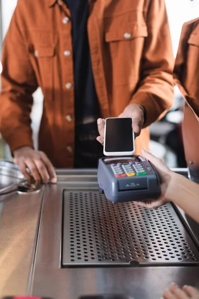 Vista recortada de la terminal de pago de tenencia de barista cerca del cliente afroamericano con teléfono inteligente en la cafetería - foto de stock