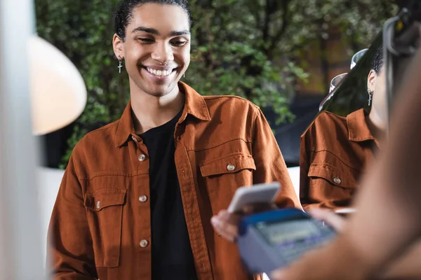Sonriente cliente afroamericano sosteniendo teléfono celular cerca de barista borrosa con terminal de pago en la cafetería - foto de stock
