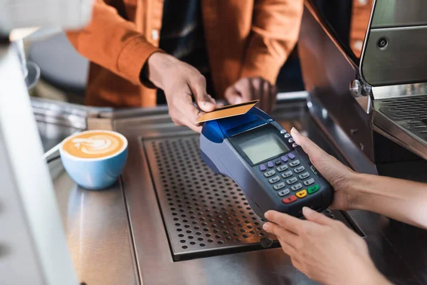 Vista recortada de la terminal de pago de tenencia de barista cerca del cliente afroamericano y café en la cafetería - foto de stock