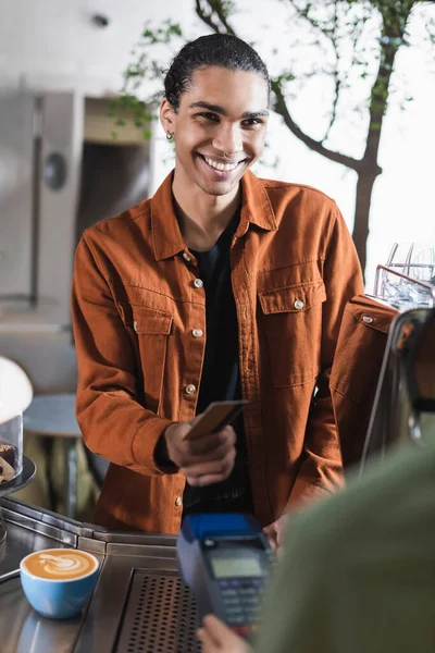 Sonriente cliente afroamericano con tarjeta de crédito cerca de barista borrosa con terminal de pago y café en la cafetería - foto de stock