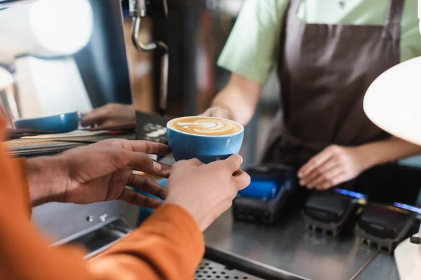 Ausgeschnittene Ansicht eines afrikanisch-amerikanischen Mannes mit Kaffee in der Nähe verschwommener Barista im Café — Stockfoto