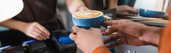 Cropped view of african american customer holding coffee cup near blurred barista in cafe, banner — Stock Photo