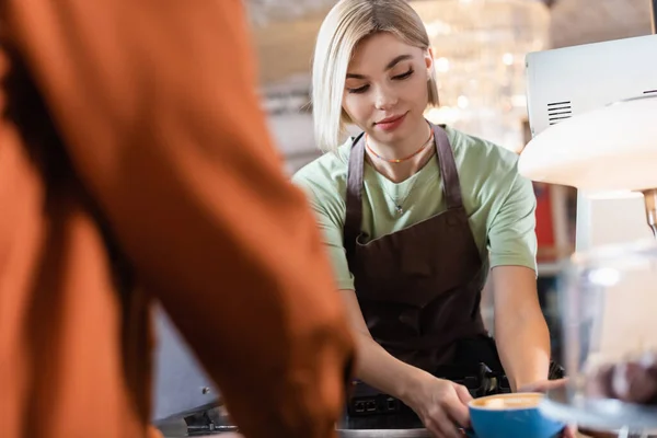 Jovem barista segurando xícara de café perto de cliente desfocado no café — Fotografia de Stock