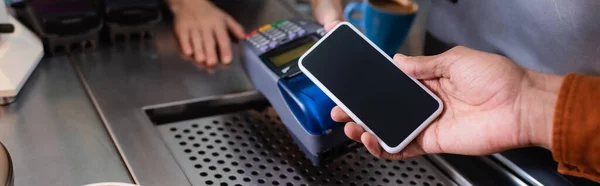 Vista recortada del hombre afroamericano que paga con teléfono celular cerca de la terminal de pago en la cafetería, pancarta - foto de stock