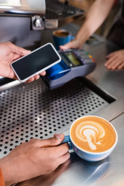 Vista recortada del hombre afroamericano sosteniendo café y teléfono inteligente cerca de barista con terminal de pago en la cafetería - foto de stock