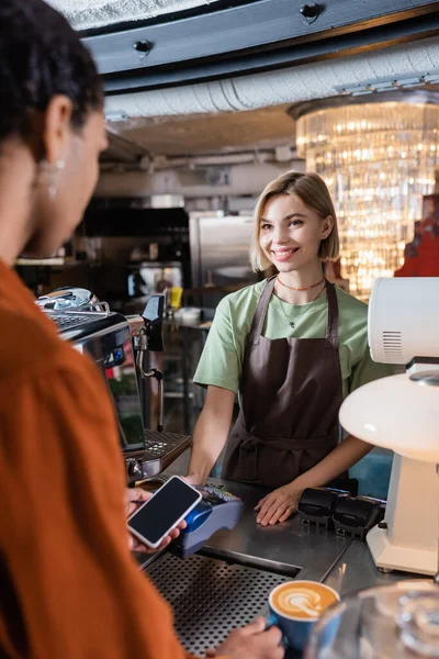 Sorridente barista con terminale di pagamento vicino al cliente afroamericano con smartphone e caffè nel caffè — Foto stock