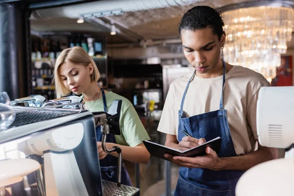 Barista afroamericano escribiendo en un cuaderno cerca de colega y máquina de café en la cafetería - foto de stock