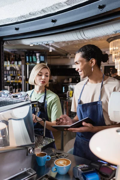 Barista afroamericano sorridente che tiene il taccuino vicino a collega, macchina da caffè e caffè in caffè — Foto stock