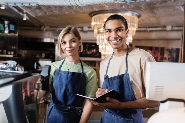 Baristi multietnici positivi con notebook che guardano la macchina fotografica in caffè — Foto stock