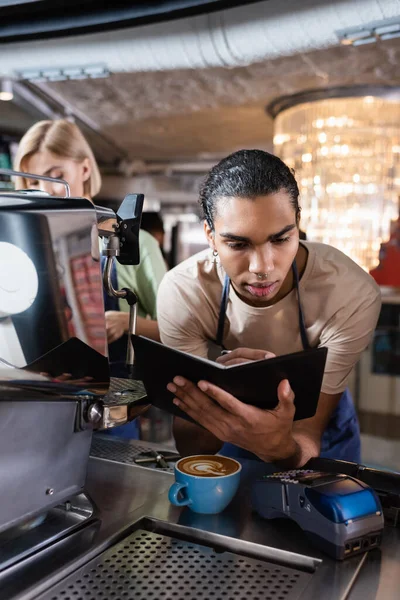 Африканський барист, який пише на записнику біля кави і платіжного терміналу в кафе. — стокове фото