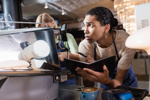Африканский американец-бариста пишет на ноутбуке возле кофеварки и платежных терминалов в кафе — стоковое фото