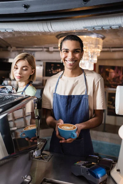 Positiver afrikanisch-amerikanischer Barista hält Tasse Kaffee in der Nähe von Kollegen und Zahlungsterminals im Café — Stockfoto