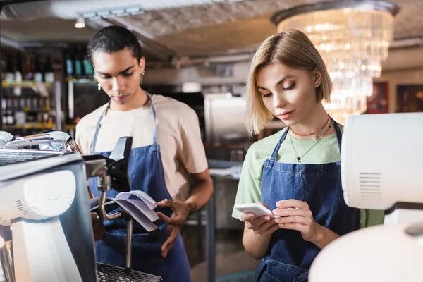 Junger Barista mit Smartphone neben afrikanisch-amerikanischem Kollegen mit Notizbuch im Café — Stockfoto