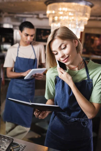 Smiling barista talking on smartphone and looking at notebook in cafe — Stock Photo