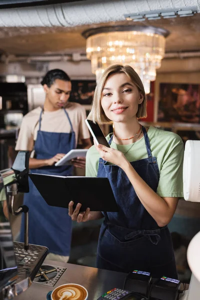 Barista souriant tenant téléphone portable et ordinateur portable près du café et des terminaux de paiement dans le café — Photo de stock