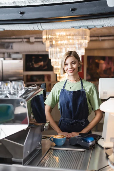 Barista sonriente mirando la cámara cerca de las terminales de café y pago en la cafetería - foto de stock