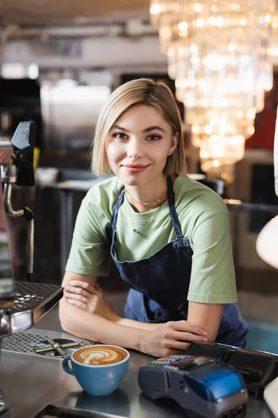 Blonde barista looking at camera near payment terminal and coffee cup in cafe — Stock Photo