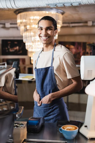 Sonriente barista afroamericano mirando a la cámara cerca de terminales de pago y café en la cafetería - foto de stock