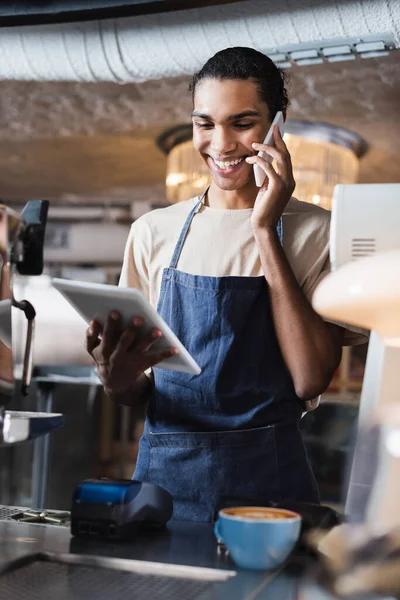 Sorrindo barista afro-americano falando no smartphone e olhando para tablet digital no café — Fotografia de Stock