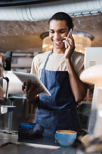 Lächelnder afrikanisch-amerikanischer Barista im Gespräch auf Smartphone und Blick auf digitales Tablet im Café — Stockfoto