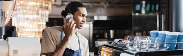 Sonriente barista afroamericano hablando en el teléfono inteligente cerca de la máquina de café en la cafetería, pancarta - foto de stock