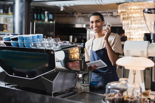 Sonriente africano americano barista dispositivos de retención cerca de la máquina de café en la cafetería - foto de stock