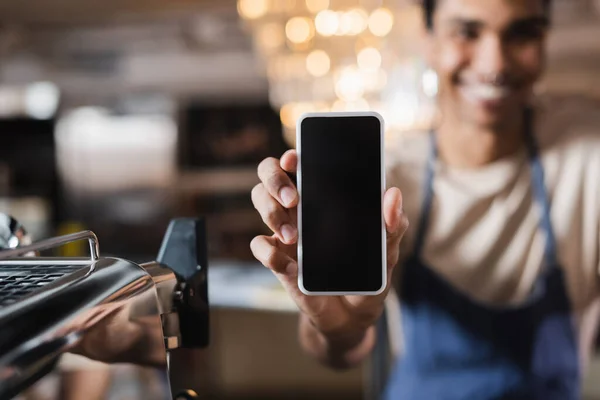 Smartphone con pantalla en blanco en la mano del borroso barista afroamericano en la cafetería - foto de stock