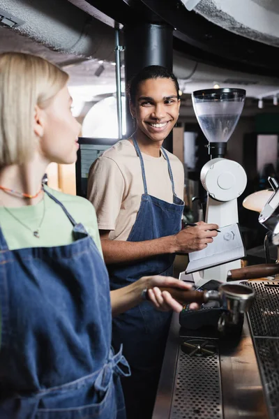 Lächelnder afrikanisch-amerikanischer Barista schreibt auf Notizbuch neben Kollege mit Kaffeeportafilter im Café — Stockfoto