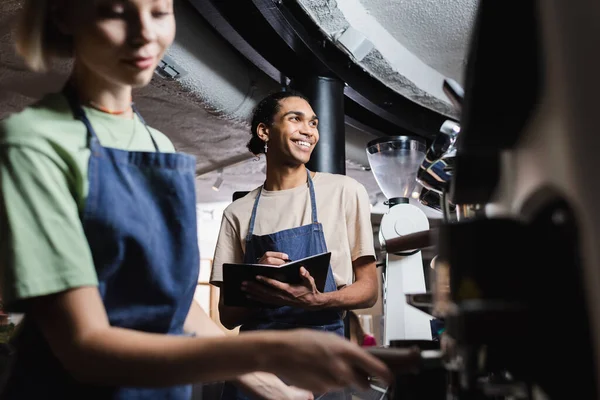 Sorrindo barista americano africano em avental escrevendo no notebook perto colega borrado e máquina de café no café — Fotografia de Stock