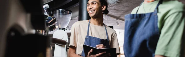 Cheerful african american barista writing on notebook near blurred colleague in cafe, banner — Stock Photo