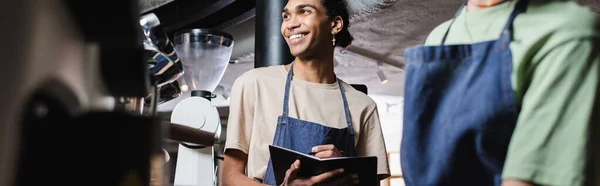 Alegre barista afroamericano escrito en un cuaderno cerca de colega borroso en la cafetería, pancarta - foto de stock