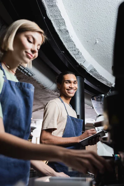 Sorrindo Africano americano barista escrevendo no notebook perto colega borrado e máquina de café no café — Fotografia de Stock