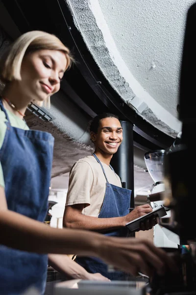 Barista afroamericano sonriente escribiendo en un cuaderno cerca de un colega borroso y una máquina de café en la cafetería - foto de stock