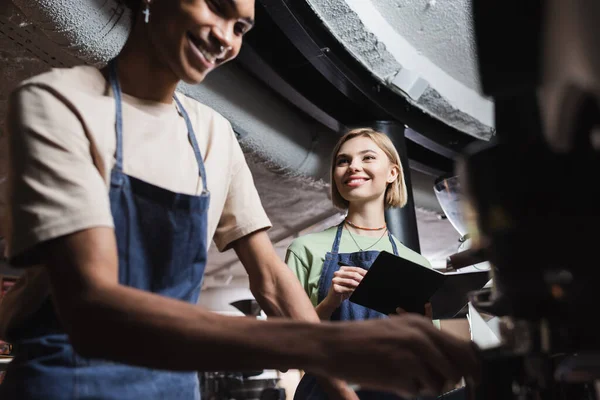 Low angle view of cheerful barista holding notebook near african american colleague and coffee machine in cafe — Stock Photo