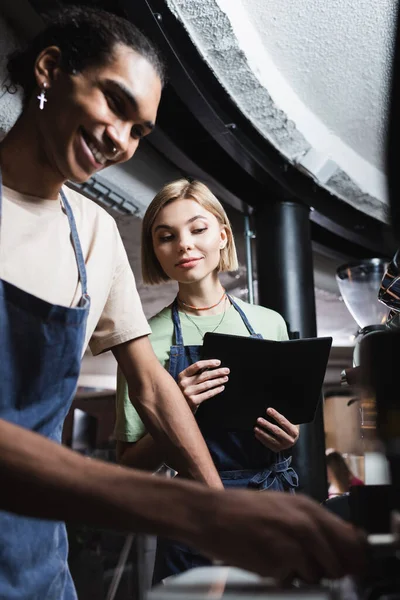 Smiling multiethnic baristas with notebook using coffee machine in cafe — Stock Photo