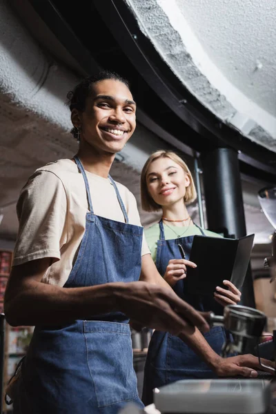 Smiling multiethnic baristas in aprons holding notebook and coffee portafilter in cafe — Stock Photo