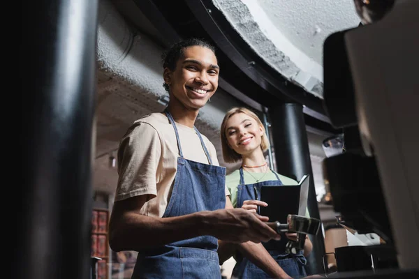 Baristas interracial positif tenant portafilter café et ordinateur portable dans le café — Photo de stock