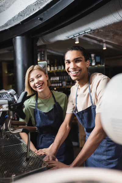 Alegre interracial baristas mirando la cámara cerca de máquina de café en la cafetería - foto de stock