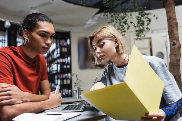 Young woman pointing at paper folder near african american freelancer and devices in cafe — Stock Photo