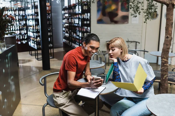 Sonriente freelancer afroamericano sosteniendo teléfono inteligente cerca de amigo y portátil en la cafetería - foto de stock