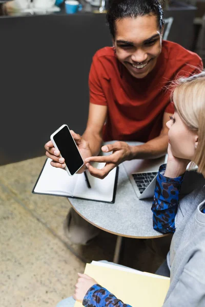 Sorridente uomo africano americano in possesso di smartphone vicino freelance in caffè — Foto stock