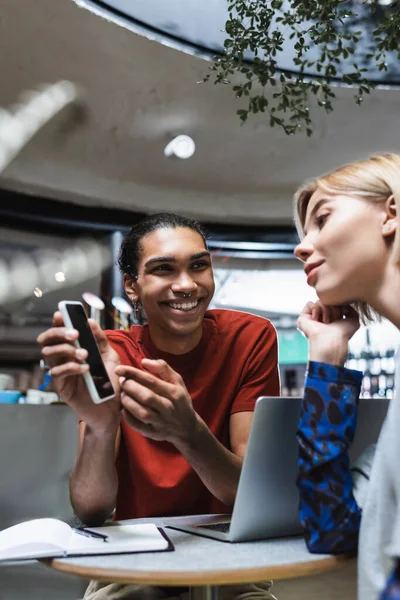 Freelancer afro-americano positivo apontando para celular perto de amigo e laptop no café — Fotografia de Stock