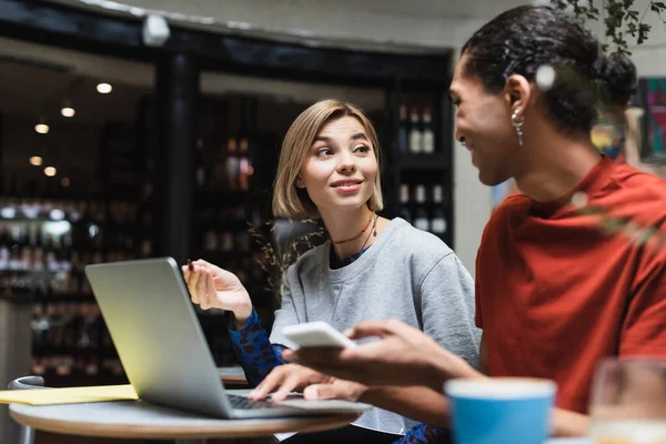 Lächelnde Freiberuflerin im Gespräch mit afrikanisch-amerikanischem Freund in der Nähe von Geräten im Café — Stockfoto